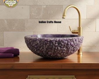 Puede incluir: Un lavabo de piedra púrpura con un exterior texturizado se encuentra sobre una encimera de madera. Un grifo de color dorado está posicionado a la derecha del lavabo. El texto "Indian Crafts House" es visible en el lavabo y en una placa en el mueble.