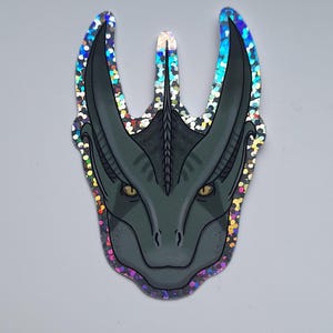 May include: A holographic sticker depicting a dragon's head in dark teal with yellow eyes and intricate details. The sticker has iridescent edges that create a shimmering effect. The dragon's head is the central focus, with a textured design and a unique shape.