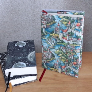 Può includere: Due libri con copertina rigida e copertine decorative. Uno presenta un gatto nero, lune e stelle su sfondo scuro. L'altro mostra un paesaggio colorato con un fiume, castelli ed edifici. Entrambi i libri hanno segnalibri a nastro.