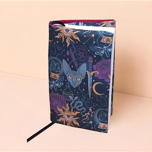 Peut inclure: Une couverture de livre en tissu avec un fond bleu marine foncé et un motif répétitif de chats sphynx, de soleils avec des yeux, de poignards, de roses violettes, de boules de cristal, de papillons de nuit, de serpents et de croissants de lune.