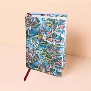 Peut inclure: Une couverture de livre rectangulaire présentant une illustration détaillée d'un paysage fantastique avec de multiples châteaux, des tours et des rivières bleues sinueuses. Le motif inclut des bâtiments aux toits rouges et un marque-page ruban rouge.