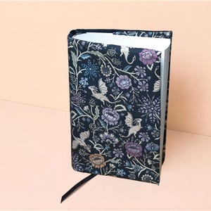 Peut inclure: Un livre à couverture rigide avec une reliure en tissu bleu marine foncé orné d'un motif répétitif de dragons ailés clairs, de fleurs violettes et bleues, et de feuillage vert. Le livre présente des pages blanches et un marque-page ruban noir.