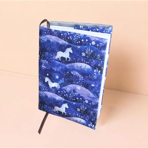 Può includere: Una copertina per libro in tessuto con un motivo blu scuro raffigurante unicorni bianchi, colline ondulate ed elementi floreali. La copertina avvolge un libro dalle pagine bianche e include un sottile segnalibro a nastro marrone.