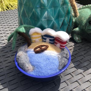 May include: A felted wool miniature beach scene in a blue bowl. The scene features a blue ocean, sand, a palm tree, and three small huts with white fluffy roofs. A pretzel sits on the sand.