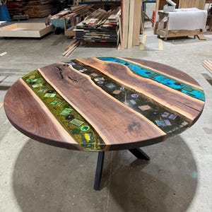 May include: Round wooden table with a dark walnut finish. The tabletop features inlaid epoxy resin in shades of green and blue, creating a river-like design. The table is supported by a black metal base.