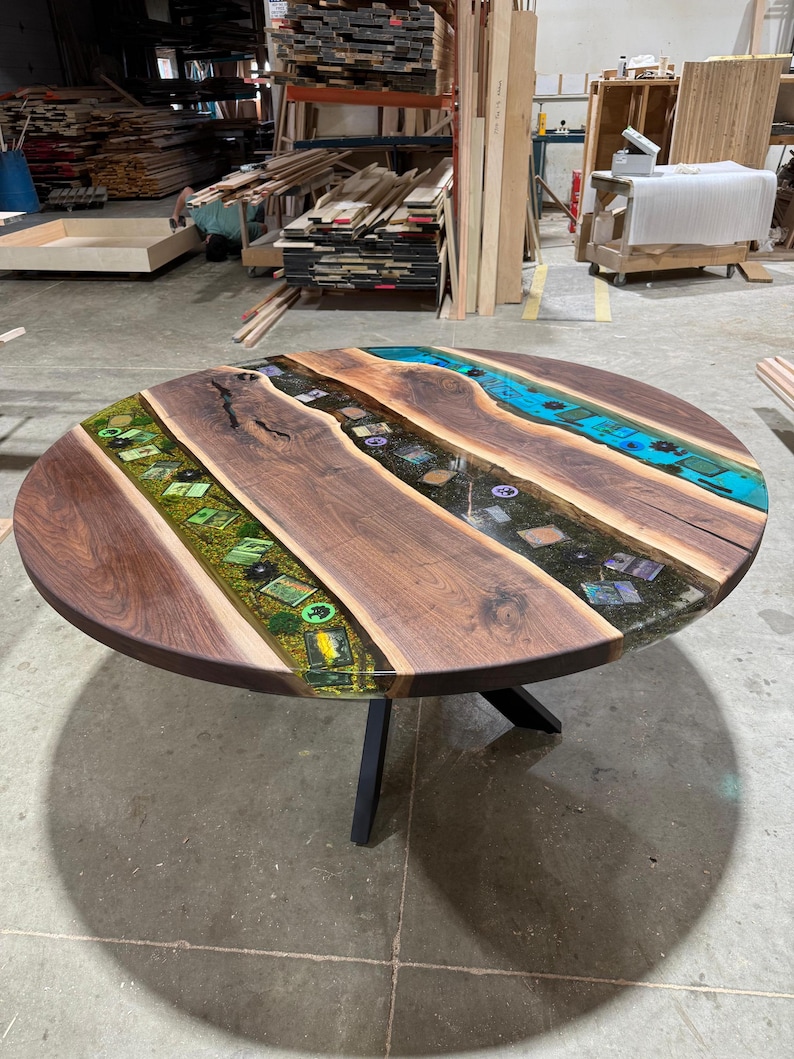 May include: Round wooden table with a dark walnut finish. The tabletop features inlaid epoxy resin in shades of green and blue, creating a river-like design. The table is supported by a black metal base.