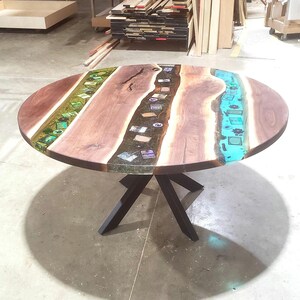 May include: A round wooden table with a unique design. The tabletop features a dark brown wood grain with sections of green, black, and turquoise resin. The table is supported by a black metal base.