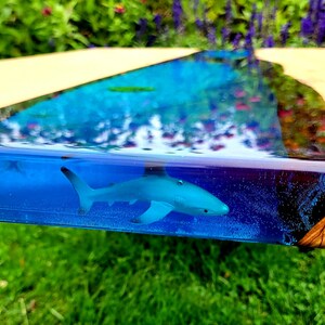 May include: A blue resin table top with a blue shark figurine embedded in the resin. The resin is clear and has a rippled effect, giving the appearance of water. The table top is set against a blurred background of green grass.