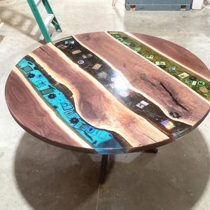 May include: A round wooden table with a dark brown finish and a unique design. The table features inlaid sections of blue, black, and green resin, each containing small decorative elements. The table is supported by a black metal base.