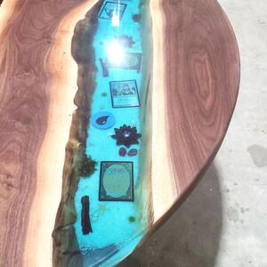 May include: Round wooden table with a river of blue resin running through the center. The river contains playing cards, a black flower, and other decorative elements. The table has a natural wood grain.