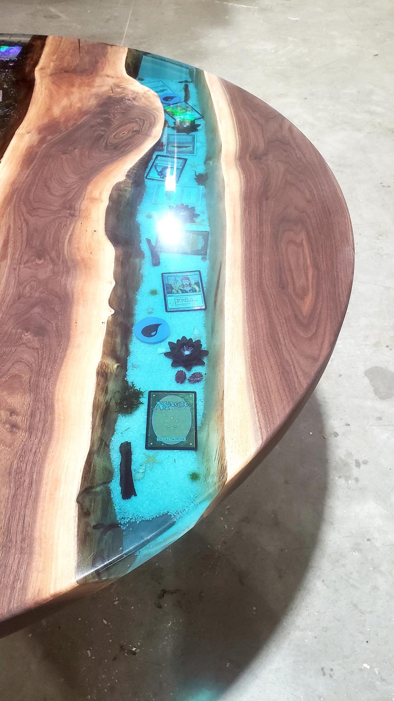 May include: Round wooden table with a river of blue resin running through the center. The river contains playing cards, a black flower, and other decorative elements. The table has a natural wood grain.