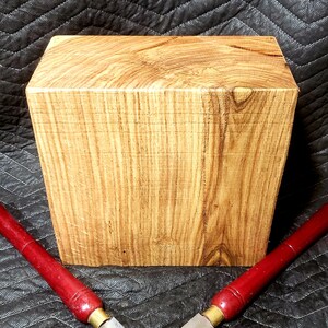 May include: A rectangular block of wood with a natural wood grain finish. The block is sitting on a black quilted surface. Two red-handled wood carving tools are crossed in front of the block.