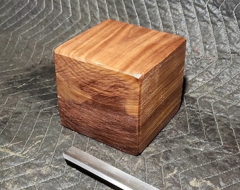 May include: A square block of wood, likely walnut, with a smooth, polished finish. The wood has a rich brown color with darker streaks throughout. Three wood carving tools are visible in the foreground.