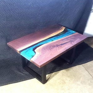 May include: A rectangular wooden coffee table with a river of turquoise resin running through the center. The wood has a reddish-brown hue, and the table is supported by black metal legs. The table measures approximately 48 inches long and 24 inches wide.