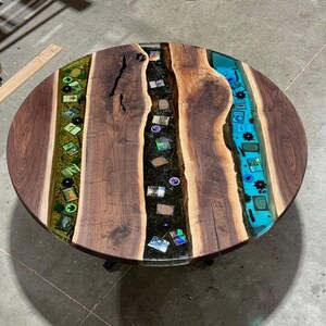 May include: Round wooden coffee table with a unique design. The table features a dark walnut wood top with a river of blue and green epoxy resin. Embedded within the resin are small decorative items. The table is supported by black metal legs.