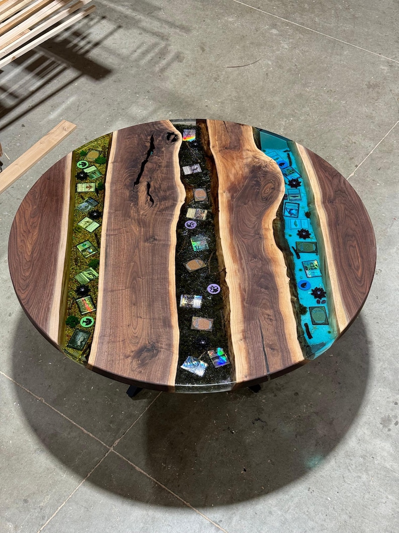 May include: Round wooden coffee table with a unique design. The table features a dark walnut wood top with a river of blue and green epoxy resin. Embedded within the resin are small decorative items. The table is supported by black metal legs.