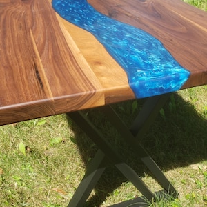 May include: A handcrafted wooden table with a vibrant blue epoxy resin river design. The table features a dark wood grain and sits on a black metal base. The table's dimensions are approximately 60 inches long and 30 inches wide.