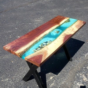 May include: A wooden table with a river of blue epoxy resin and small stones embedded in the resin. The table has a black X-shaped base.
