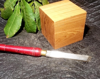 May include: A wooden cube with visible grain, a red-handled chisel, and green leaves rest on a textured black surface. The chisel has a silver blade and brass ferrule. The cube is a light brown color.