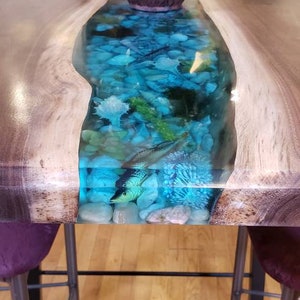 May include: A wooden table with a blue resin river running through the center. The river is filled with small white and blue stones and a few small fish. The table is sitting on a wooden floor.