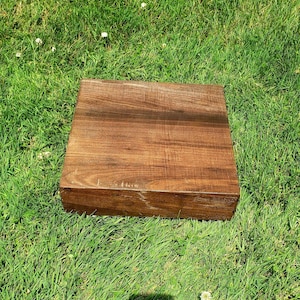 May include: A square, wooden box with a rich, brown finish. The box is made of wood and has a natural grain pattern. The box is sitting on a bed of green grass.