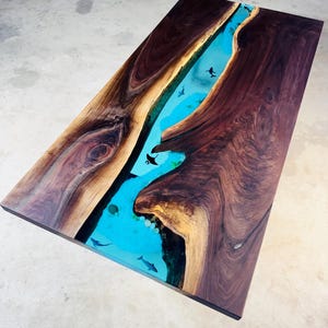 May include: A rectangular wooden coffee table with a river-like design. The table features a dark brown wood frame with a turquoise blue epoxy resin center, complete with small fish illustrations. The table measures approximately 60 inches long and 30 inches wide.