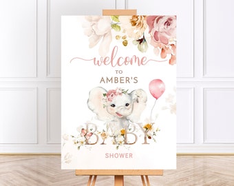 May include: A welcome sign for a baby shower with a pink and white floral design. The sign features a cute cartoon elephant with a pink flower crown and a pink balloon. The text on the sign reads "Welcome to Amber's Baby Shower".