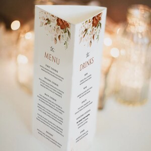 Op de afbeelding: Een drievoudig gevouwen huwelijksmenukaart met een bloemenontwerp in tinten wit, bruin en oranje. Het menu bevat een voorgerecht, een hoofdgerecht, een hoofdgerecht, een dessert en drankjes. De tekst "the MENU" en "the DRINKS" is in bruin gedrukt.