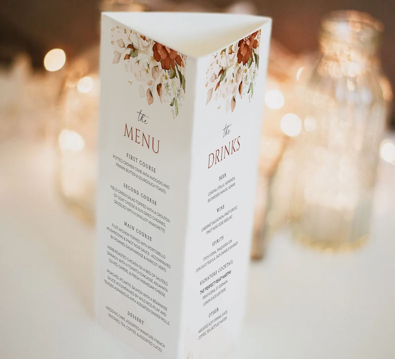 Op de afbeelding: Een drievoudig gevouwen huwelijksmenukaart met een bloemenontwerp in tinten wit, bruin en oranje. Het menu bevat een voorgerecht, een hoofdgerecht, een hoofdgerecht, een dessert en drankjes. De tekst "the MENU" en "the DRINKS" is in bruin gedrukt.