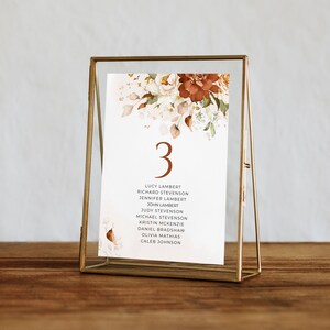 May include: A gold-framed table number card with the number "3" in a brown font. The card features a watercolour floral design with white, peach, and rust-coloured flowers and leaves. The card lists the names of guests for table number 3: Lucy Lambert, Richard Stevenson, Jennifer Lambert, John Lambert, Judy Stevenson, Michael Stevenson, Kristin McKenzie, Daniel Bradshaw, Olivia Mathias, and Caleb Johnson.