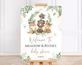 May include: Welcome to Meadow & River's baby shower sign with a woodland theme. The sign features a brown bear, a fox, a deer, and a raccoon surrounded by green leaves and flowers. The text on the sign reads "Welcome to Meadow & River's baby shower 11.06.23".