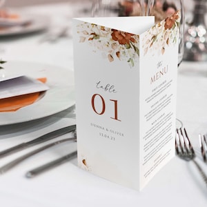 Op de afbeelding: Een witte tafelnummerkaart voor een bruiloft met een bloemenontwerp in tinten bruin en wit. De kaart heeft de tekst "tafel 01" in bruin en "SIENNA & OLIVER 13.04.23" in zwart. De kaart ligt op een wit tafelkleed met bestek en een glas.