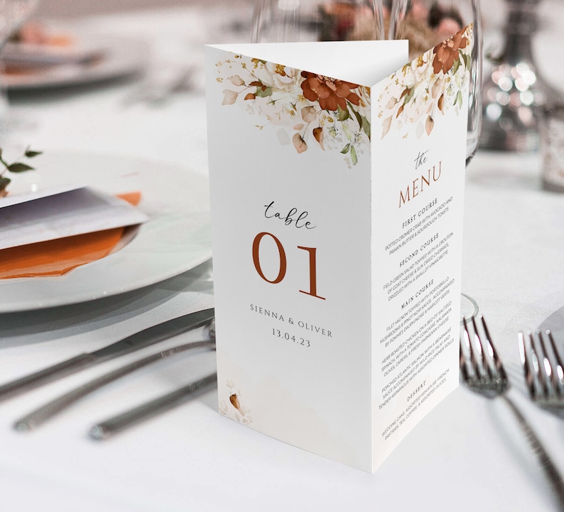 Op de afbeelding: Een witte tafelnummerkaart voor een bruiloft met een bloemenontwerp in tinten bruin en wit. De kaart heeft de tekst "tafel 01" in bruin en "SIENNA & OLIVER 13.04.23" in zwart. De kaart ligt op een wit tafelkleed met bestek en een glas.