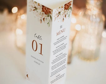 May include: A white wedding table number card with a floral design in shades of orange and white. The card has the text "table 01" in brown text and the names "SIENNA & OLIVER" and the date "13.04.23" in smaller brown text. The menu is printed on the back of the card.