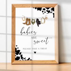 May include: A white sign with black text and a cow print border. The text reads "babies are sweet" and "please take a treat". There are illustrations of cowboy boots, a bandana, a hat, a horseshoe, and a star hanging from a string.
