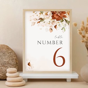 May include: A white table number card with a floral design in shades of brown, orange, and white. The text "table number 6" is printed in brown.