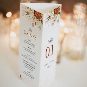 Op de afbeelding: Een witte tafelnummerkaart met een bloemenontwerp en de tekst "the DRINKS" in bruine letters. Het tafelnummer is "01" in een bruin lettertype. De kaart bevat ook een lijst met drankjes, waaronder bier, wijn, sterke drank, een signature cocktail en andere opties. Onderaan de kaart staat "SIENNA & OLIVER 13.04.23".