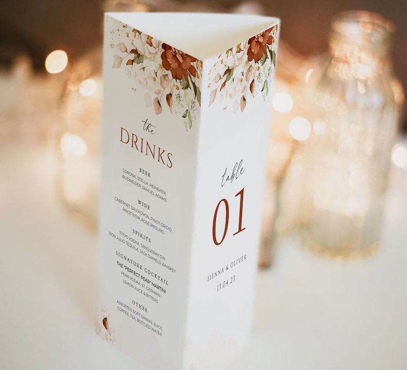 Op de afbeelding: Een witte tafelnummerkaart met een bloemenontwerp en de tekst "the DRINKS" in bruine letters. Het tafelnummer is "01" in een bruin lettertype. De kaart bevat ook een lijst met drankjes, waaronder bier, wijn, sterke drank, een signature cocktail en andere opties. Onderaan de kaart staat "SIENNA & OLIVER 13.04.23".