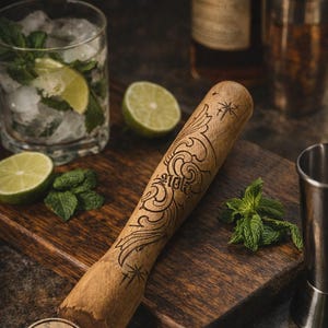 May include: A handmade wooden muddler with etched patterns, labelled "The Wayfinder Muddler." The image shows a cocktail glass with ice and mint, lime slices, and a metal shaker. The text reads "Designed & Burned by the Artist."