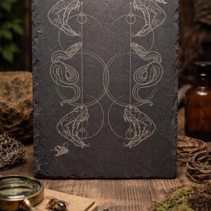 May include: A rectangular black slate board featuring an etched design of four snakes facing each other. The snakes have open mouths and detailed patterns, arranged in pairs. The design is in a light beige colour.