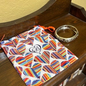 May include: A silver bangle bracelet with an ornate design, in a white drawstring bag with a colourful heart pattern and the letter "B" in a heart.