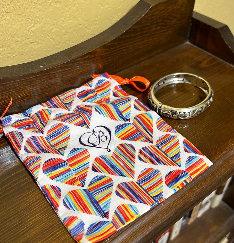 May include: A silver bangle bracelet with an ornate design, in a white drawstring bag with a colourful heart pattern and the letter "B" in a heart.