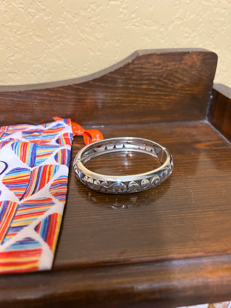 May include: A silver bangle bracelet with an intricate floral design. The bracelet is open and has a hinged clasp.