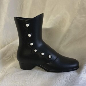 May include: A matte black decorative boot figurine featuring a side closure with five small, raised white circular buttons arranged along a scalloped edge. The boot has a low heel, a rounded toe, and a smooth finish, standing upright as a display piece.