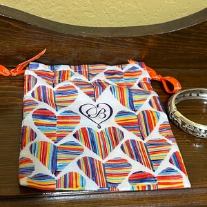 May include: A silver bangle bracelet with an intricate floral design. The bracelet is in a small, white drawstring bag with a colourful heart pattern and the letter "B" in a heart.