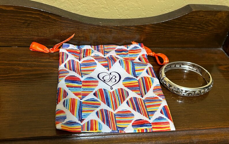 May include: A silver bangle bracelet with an intricate floral design. The bracelet is in a small, white drawstring bag with a colourful heart pattern and the letter "B" in a heart.