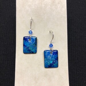May include: A pair of blue and green enamel earrings with silver hooks and blue crystal accents. The earrings are square shaped.