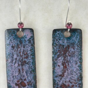 Puede incluir: Un par de pendientes rectangulares con un diseño marmoleado azul, morado y blanco. Los pendientes tienen un gancho plateado con una pequeña perla morada en la parte superior.