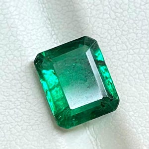 May include: A rectangular emerald gemstone, displaying a rich green colour. The gem is cut with facets, revealing its clarity and sparkle. The emerald is set against a white backdrop, accentuating its colour and form. A gemstone representing luxury and beauty.