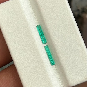 Op de afbeelding: Twee levendige, smaragdgroene edelsteenkristallen worden getoond tegen een witte, getextureerde ondergrond. De kristallen zijn rechthoekig met een enigszins doorschijnend uiterlijk, wat hun interne structuur laat zien. De edelstenen zijn diagonaal geplaatst.